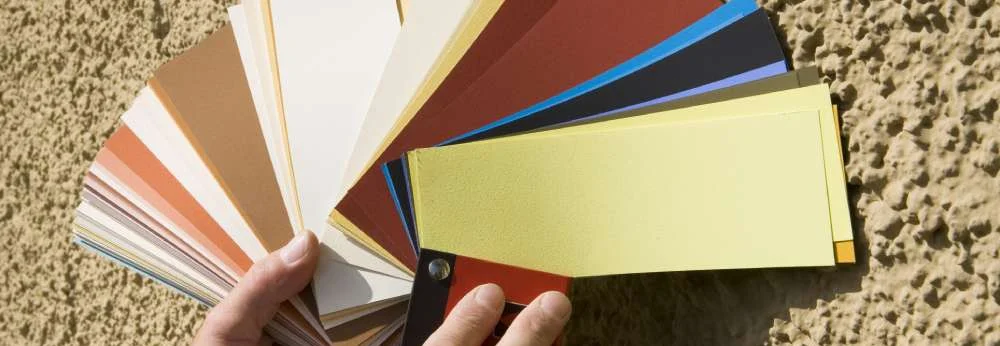 Eine Person hält einen Farbtonfächer an eine verputzte Hauswand.